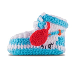 May include: A handmade crochet baby bootie in the style of a high-top sneaker. The bootie features white and light blue yarn with a red accent and the word "AIR" in black. The design is a unique and playful take on a classic shoe.