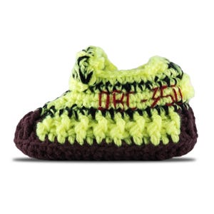 May include: A handmade crochet baby shoe in a sneaker style. The shoe features a yellow and black upper with a brown sole. The text "DRC-350" is embroidered in red on the side. The shoe is designed to resemble a popular sneaker.