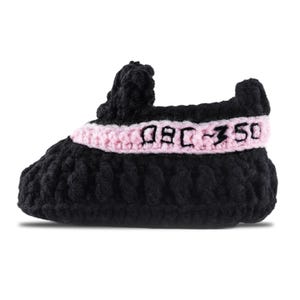 May include: A black and pink crocheted baby shoe. The shoe features a black body with a pink band across the middle. The text "08C-350" is embroidered on the pink band. The shoe is designed to resemble a popular sneaker style.