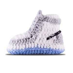 May include: A handmade crochet baby sneaker in white, gray, and blue. The sneaker features a high-top design with a blue sole, gray accents, and a black stitched detail. The yarn is thick and the stitches are visible.
