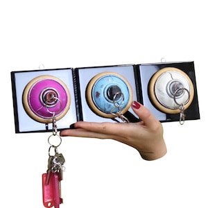 Op de afbeelding: Een sleutelhouder met drie ronde sleutelhaken in roze, blauw en zilver. Elke haak is geplaatst in een vierkant zwart frame met een houten rand. Een set sleutels hangt aan de roze sleutelhaken.