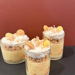 Może przedstawiać: Trzy szklane świece o tematyce chleba bananowego. Każda świeca ma żółtą podstawę, zwieńczoną białą bitą woskiem, płatkami zbożowymi i woskowymi akcentami w kształcie banana. Etykieta głosi "Nana's Banana Bread".