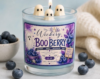 May include: A light blue soy candle in a glass jar with three ghost-shaped wax toppers and a lit wick. The label reads The Wickery BOO BERRY, featuring illustrations of blueberries, a ghost, and text stating 10 OZ, blueberry, vanilla, marshmallow, sugar.