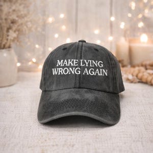 May include: A dark gray baseball cap with the embroidered text "MAKE LYING WRONG AGAIN" in white. The cap has a curved brim and a fabric strap closure. The background includes soft lighting and decorative elements.