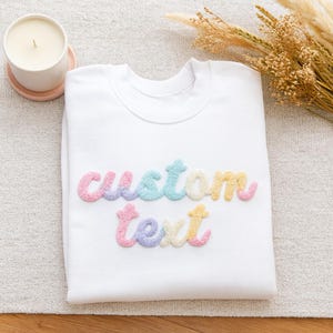 May include: A white sweatshirt with the words "custom text" in colorful, textured letters. The letters are in pastel shades of pink, blue, yellow, and purple. A lit candle in a white ceramic holder sits on a pink coaster.