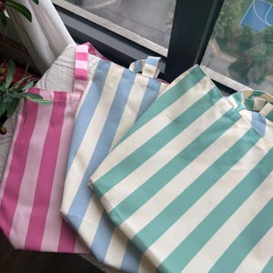 May include: Three tote bags in pink, light blue, and teal, each with wide white stripes. The bags have matching striped handles and are made of a sturdy fabric. The bags are ideal for carrying groceries, books, or other items.