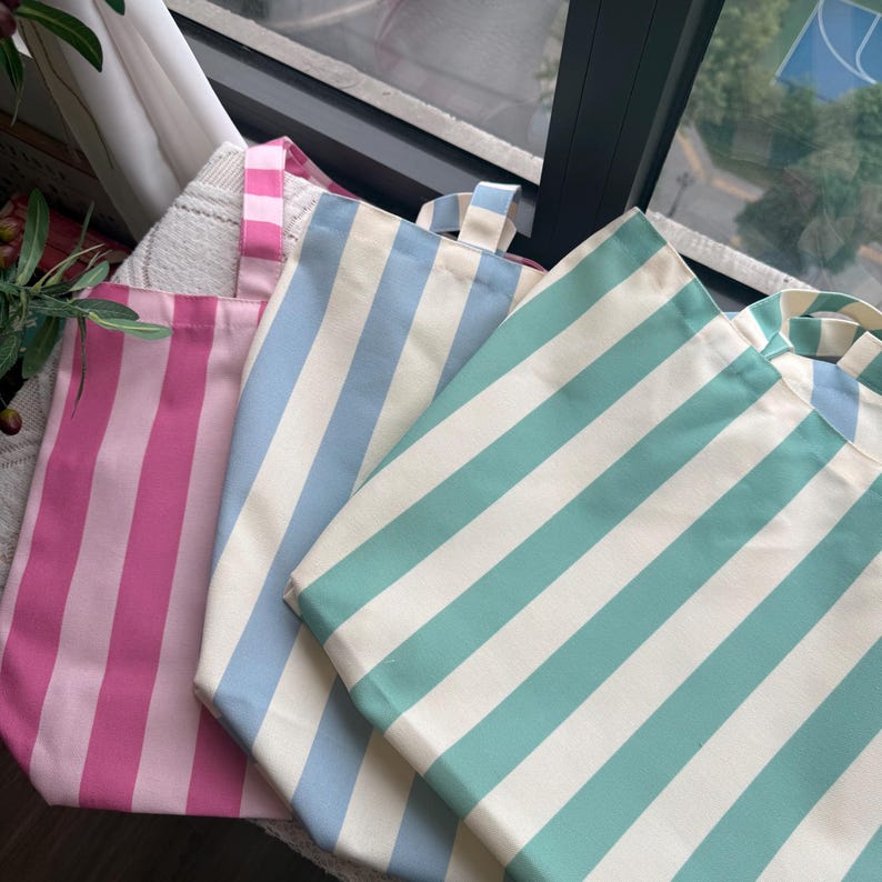 May include: Three tote bags in pink, light blue, and teal, each with wide white stripes. The bags have matching striped handles and are made of a sturdy fabric. The bags are ideal for carrying groceries, books, or other items.