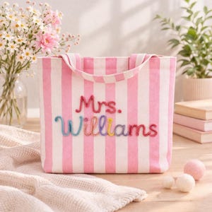 May include: A pink and white striped tote bag with the embroidered text "Mrs. Williams" in colorful thread. The bag has two handles and is sitting on a light-colored surface. Decorative elements include flowers and small felt balls.