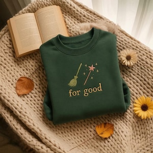 May include: A dark green sweatshirt with the embroidered words "for good" below a broom and a magic wand. The sweatshirt is on a beige knitted blanket with a book and fall leaves.