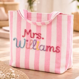 May include: A pink and white striped tote bag with the embroidered name "Mrs. Williams" in colorful thread. The bag has matching pink handles and is made of a sturdy fabric. The bag is a perfect accessory for carrying essentials.