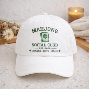 May include: A white baseball cap featuring dark green embroidered text that reads Mahjong Social Club, Est 2026, Cracks, Dots, Bams, and a central light green Mahjong tile icon. The cap has a structured crown and a curved brim.