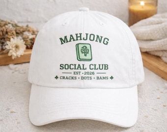 Op de afbeelding: Witte baseballcap met groene tekst "MAHJONG SOCIAL CLUB". De cap heeft een groen embleem en de tekst "EST. 2026" met "CRACKS • DOTS • BAMS" eronder. Klassieke stijl met een gebogen klep.