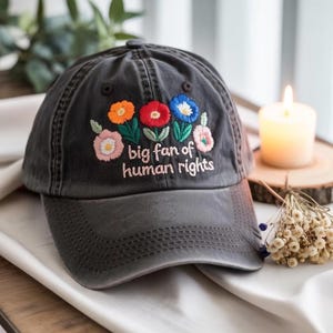 Op de afbeelding: Een donkergrijze baseballcap met geborduurde kleurrijke bloemen en de tekst "big fan of human rights". De bloemen zijn in tinten oranje, rood, blauw en roze. De cap ligt op een witte ondergrond.