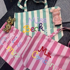 May include: Three striped tote bags with embroidered names: "Miss Becky", "Miss Camp", and "Ms. Concannon". The bags feature vertical stripes in mint green and pink, with colorful lettering. A pink bear figurine and a gold bear figurine are also visible.