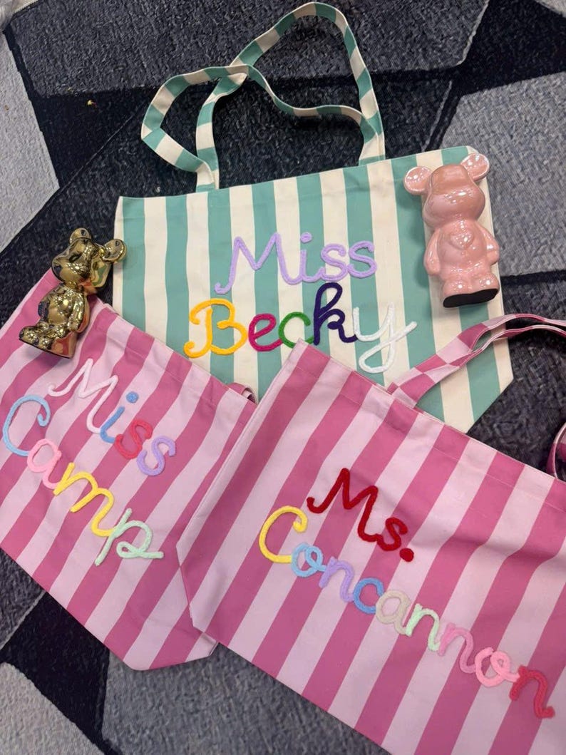 May include: Three striped tote bags with embroidered names: "Miss Becky", "Miss Camp", and "Ms. Concannon". The bags feature vertical stripes in mint green and pink, with colorful lettering. A pink bear figurine and a gold bear figurine are also visible.