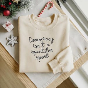 Può includere: Felpa color crema con la scritta "Democracy isn't a spectator sport" in corsivo nero. La felpa è piegata su una superficie di legno, con decorazioni natalizie tra cui un bastoncino di zucchero e un fiocco di neve.