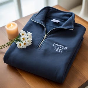 Könnte beinhalten: Ein marineblaues Sweatshirt mit Viertelreißverschluss und dem gestickten Text "CUSTOM TEXT" in Weiß. Das gefaltete Sweatshirt liegt auf einer Holzoberfläche, neben einer brennenden Kerze und einem Zweig weißer Gänseblümchen.
