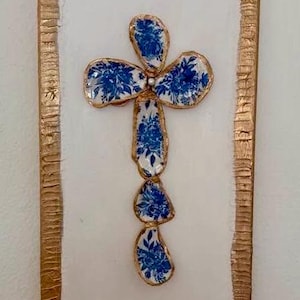 May include: A decorative cross made from blue and white floral patterned ceramic pieces, set against a white background. The cross is framed with a textured, metallic gold border, creating a striking contrast.
