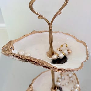 May include: Two-tiered jewelry stand with a gold-colored handle and frame. The white trays have gold-painted edges, resembling oyster shells. The top tray holds earrings and pearls, while the bottom tray has a pearl necklace.