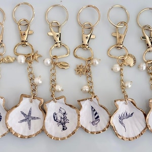May include: Six gold-toned keychains with seashell-shaped pendants. Each pendant features a different nautical design in blue, including a nautilus, starfish, lobster, sailboat, seaweed, and crab. The keychains have pearl accents.