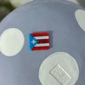 May include: A small, pixelated Puerto Rican flag in red, white, and blue is affixed to a light purple surface with white polka dots. The flag features a blue triangle with a white star and horizontal stripes.