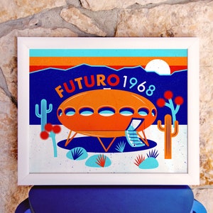 以下が含まれることがあります： 砂漠の中に「FUTURO 1968」という文字が書かれた、未来的なオレンジとブルーの家の白い額縁入りプリント。家は丸みを帯びた形をしていて、側面にドアがあります。背景にはサボテンやその他の砂漠の植物があります。