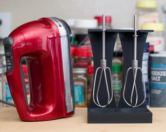 Op de afbeelding: Een rode handmixer met een zilveren accent en een zwarte standaard met twee draadgardes. De mixer heeft een snelheidsregelaar. Keukenapparaat voor bakken en koken.