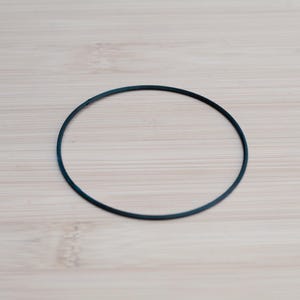 Pode incluir: Um fino anel de borracha azul-petr&oacute;leo escuro, de forma circular, repousa sobre uma superf&iacute;cie de madeira clara. A textura lisa do anel contrasta com o gr&atilde;o da madeira. O objeto est&aacute; centrado na moldura, proporcionando uma est&eacute;tica simples e minimalista.