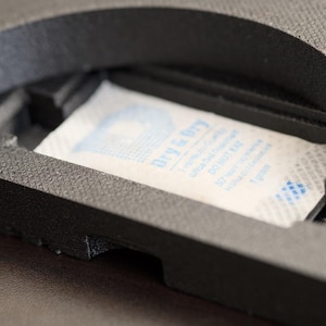 Pode incluir: Close-up de um objeto preto, texturizado e semicircular com um compartimento retangular. Dentro do compartimento h&aacute; um pacote branco com texto azul que diz "Dry & Dry". O pacote cont&eacute;m 1 grama de gel de s&iacute;lica.