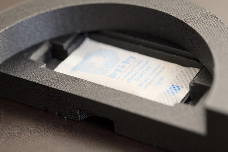 Pode incluir: Close-up de um objeto preto, texturizado e semicircular com um compartimento retangular. Dentro do compartimento h&aacute; um pacote branco com texto azul que diz "Dry & Dry". O pacote cont&eacute;m 1 grama de gel de s&iacute;lica.