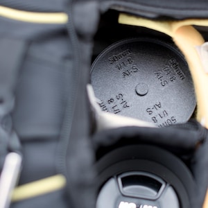 Pode incluir: Um filtro de lente preto com a inscri&ccedil;&atilde;o "50mm 1/1.8 AI-S" est&aacute; guardado dentro de uma bolsa de c&acirc;mera preta. O filtro &eacute; redondo e tem uma superf&iacute;cie texturizada. A bolsa tem detalhes amarelos.