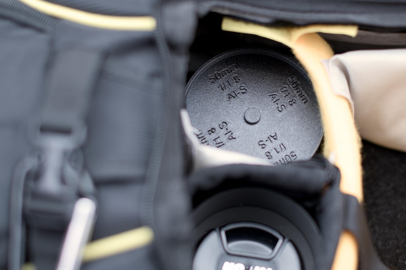 Pode incluir: Um filtro de lente preto com a inscri&ccedil;&atilde;o "50mm 1/1.8 AI-S" est&aacute; guardado dentro de uma bolsa de c&acirc;mera preta. O filtro &eacute; redondo e tem uma superf&iacute;cie texturizada. A bolsa tem detalhes amarelos.