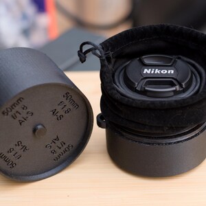 Pode incluir: Estojo preto para lente de c&acirc;mera e tampa de lente preta com o logotipo da Nikon. O estojo &eacute; cil&iacute;ndrico com o texto "50mm f/1.8 AI-S" impresso na lateral. A tampa da lente est&aacute; dentro de um saco de veludo preto.