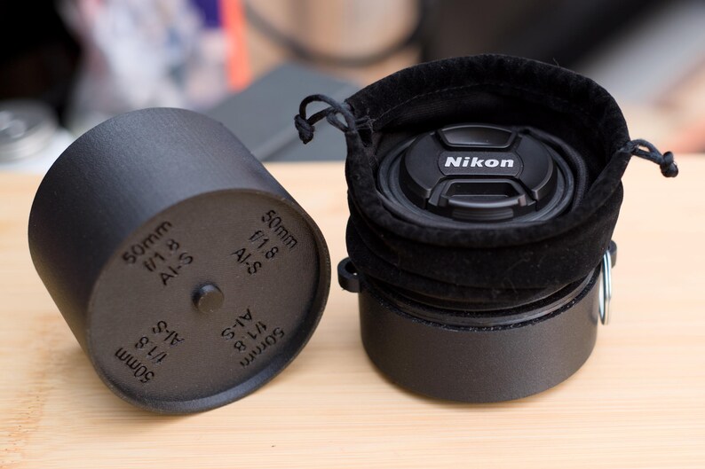 Pode incluir: Estojo preto para lente de c&acirc;mera e tampa de lente preta com o logotipo da Nikon. O estojo &eacute; cil&iacute;ndrico com o texto "50mm f/1.8 AI-S" impresso na lateral. A tampa da lente est&aacute; dentro de um saco de veludo preto.