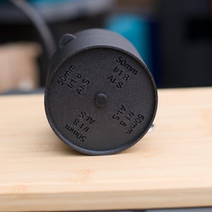 Pode incluir: Um componente de lente de c&acirc;mera cil&iacute;ndrico preto com o texto "50mm 1/1.8 AI-S" impresso na parte inferior. O componente est&aacute; sobre uma superf&iacute;cie de madeira clara. O componente da lente &eacute; de cor fosca escura.