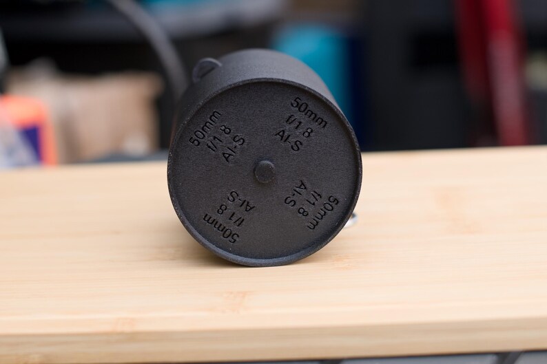 Pode incluir: Um componente de lente de c&acirc;mera cil&iacute;ndrico preto com o texto "50mm 1/1.8 AI-S" impresso na parte inferior. O componente est&aacute; sobre uma superf&iacute;cie de madeira clara. O componente da lente &eacute; de cor fosca escura.