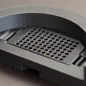 Pode incluir: Close-up de um objeto cinza escuro, semicircular, com uma superf&iacute;cie texturizada. O objeto apresenta uma se&ccedil;&atilde;o retangular com uma grade de pequenos orif&iacute;cios espa&ccedil;ados uniformemente. O design parece ser um componente ou pe&ccedil;a.