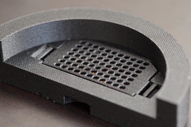 Pode incluir: Close-up de um objeto cinza escuro, semicircular, com uma superf&iacute;cie texturizada. O objeto apresenta uma se&ccedil;&atilde;o retangular com uma grade de pequenos orif&iacute;cios espa&ccedil;ados uniformemente. O design parece ser um componente ou pe&ccedil;a.