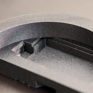 Pode incluir: Close-up de um objeto preto, semicircular, com superf&iacute;cie texturizada. O objeto tem um design em camadas com um interior rebaixado e uma borda superior arredondada. O material parece ser um pl&aacute;stico fosco ou composto.