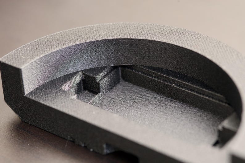 Pode incluir: Close-up de um objeto preto, semicircular, com superf&iacute;cie texturizada. O objeto tem um design em camadas com um interior rebaixado e uma borda superior arredondada. O material parece ser um pl&aacute;stico fosco ou composto.