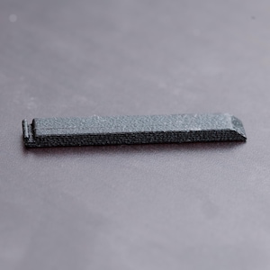 Pode incluir: Um pequeno objeto retangular cinza escuro com uma superf&iacute;cie texturizada. O objeto tem uma borda ligeiramente chanfrada em uma extremidade e uma pequena se&ccedil;&atilde;o elevada na outra. Parece ser feito de um material denso.