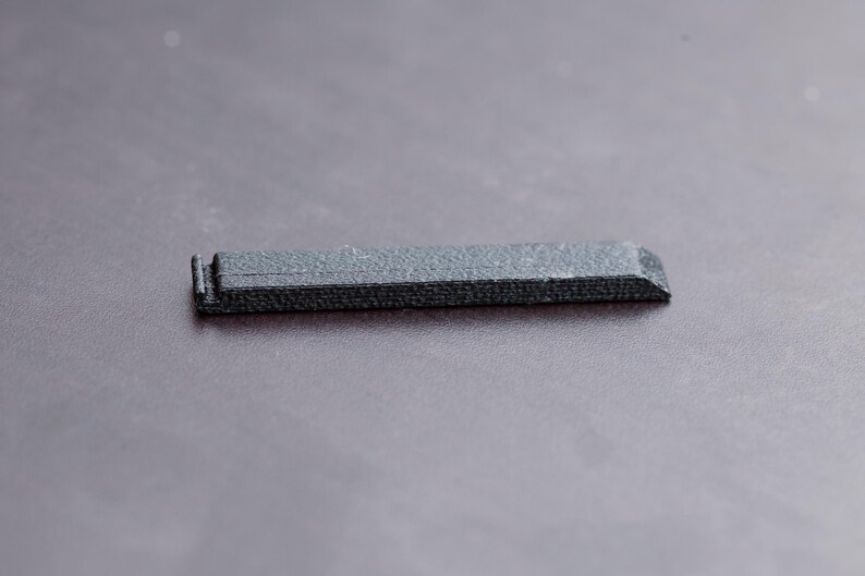 Pode incluir: Um pequeno objeto retangular cinza escuro com uma superf&iacute;cie texturizada. O objeto tem uma borda ligeiramente chanfrada em uma extremidade e uma pequena se&ccedil;&atilde;o elevada na outra. Parece ser feito de um material denso.