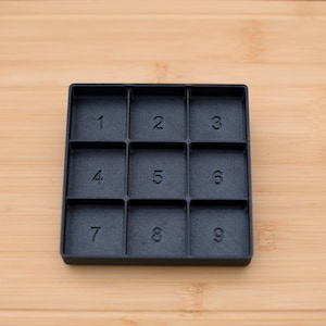 May include: A black, square organizer with nine numbered compartments. Each compartment is labeled with a number from 1 to 9. The organizer sits on a light brown wooden surface.