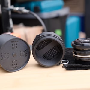 Pode incluir: Um conjunto de tampa e estojo de lente preta de 50 mm com uma lente Nikon em um pano preto. O estojo est&aacute; aberto, revelando um interior texturizado. A tampa da lente tem o texto "50mm 1:1.8 AFS" impresso.
