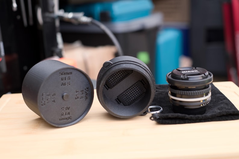 Pode incluir: Um conjunto de tampa e estojo de lente preta de 50 mm com uma lente Nikon em um pano preto. O estojo est&aacute; aberto, revelando um interior texturizado. A tampa da lente tem o texto "50mm 1:1.8 AFS" impresso.