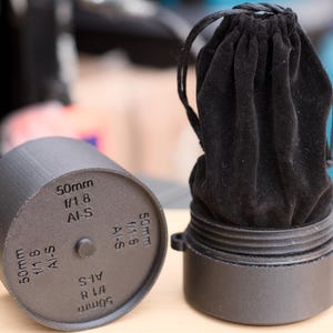 Pode incluir: Uma caixa de lente preta com uma tampa de lente cil&iacute;ndrica correspondente. A tampa &eacute; marcada com o texto "50mm f/1.8 AI-S". A caixa cont&eacute;m um saco de veludo preto com cord&atilde;o.