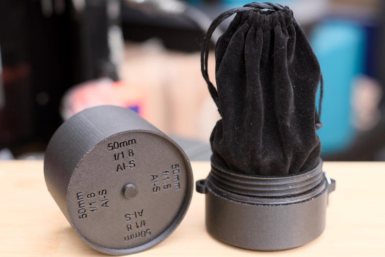 Pode incluir: Uma caixa de lente preta com uma tampa de lente cil&iacute;ndrica correspondente. A tampa &eacute; marcada com o texto "50mm f/1.8 AI-S". A caixa cont&eacute;m um saco de veludo preto com cord&atilde;o.