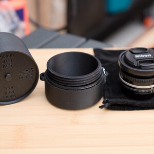Pode incluir: Lente de c&acirc;mera preta com uma tampa de lente Nikon, um estojo de lente preto e uma tampa de lente preta com texto. A lente est&aacute; em uma bolsa de veludo preta. A tampa da lente tem o texto "50mm 1/1.8 AFS".
