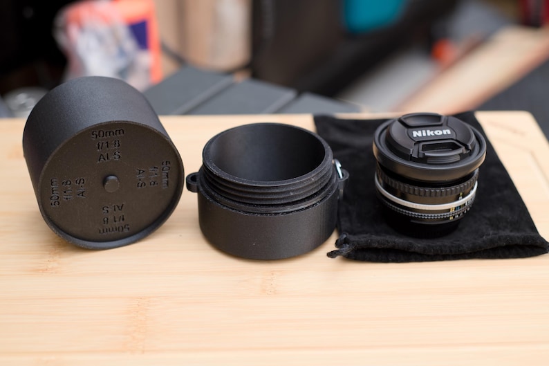 Pode incluir: Lente de c&acirc;mera preta com uma tampa de lente Nikon, um estojo de lente preto e uma tampa de lente preta com texto. A lente est&aacute; em uma bolsa de veludo preta. A tampa da lente tem o texto "50mm 1/1.8 AFS".