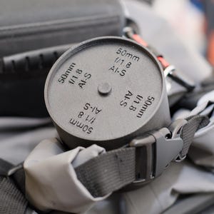 Pode incluir: Uma tampa de lente cinza com o texto "50mm f/1.8 AI-S" est&aacute; presa a uma bolsa de c&acirc;mera. A tampa da lente &eacute; cil&iacute;ndrica com um pequeno bot&atilde;o na parte superior. A bolsa tem uma al&ccedil;a cinza e uma fivela preta.